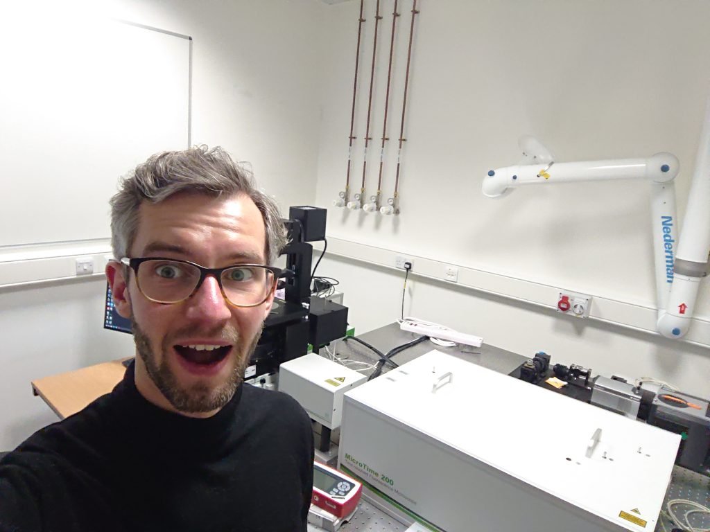 Simon beside a spectroscopy and microscopy setup in the laboratory